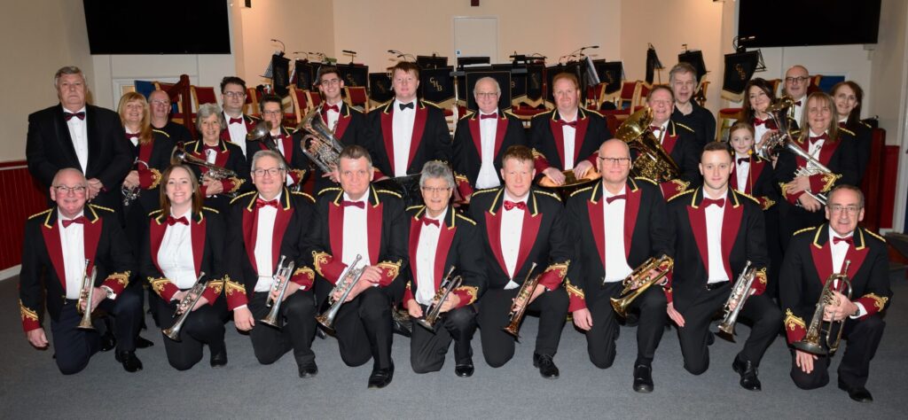 Members of Chiltern Hills Brass in black, red and gold uniforms, in two rows, smiling at the camera.